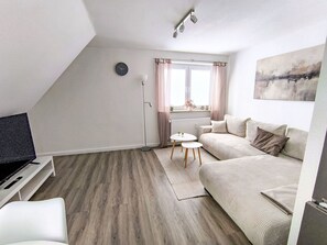 Ein modernes Wohnzimmer mit einer Couch, einem kleinen Tisch, einem Fernseher auf einem Schrank und einem Fenster mit Vorhängen.
