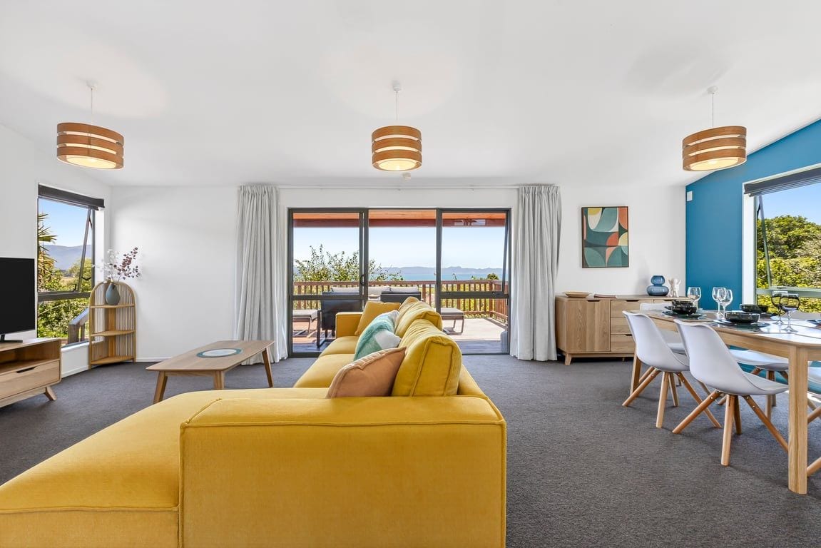 Una sala de estar moderna con un sofá amarillo, una mesa de comedor y sillas, ventanas amplias con vistas, e iluminación colgante.