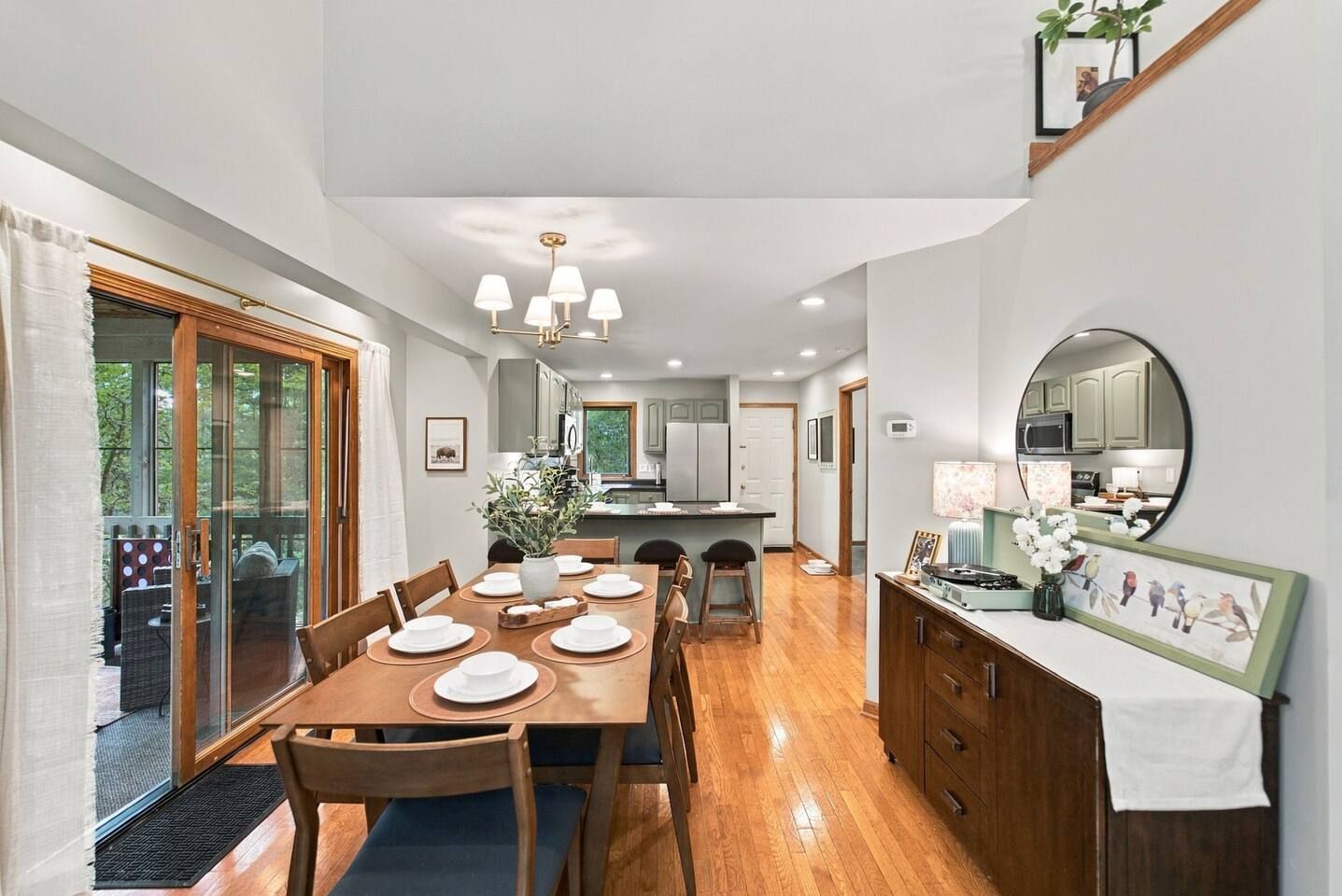 A dining area with a wooden table set for four, a kitchen in the background, and a large mirror on the wall.