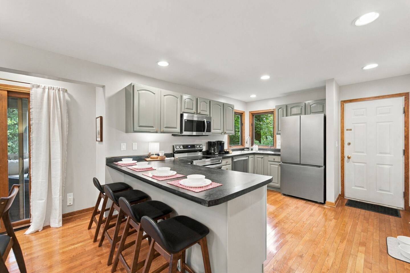 A modern kitchen with grey cabinets, stainless steel appliances, a central island with bar stools, and a large window providing natural light.