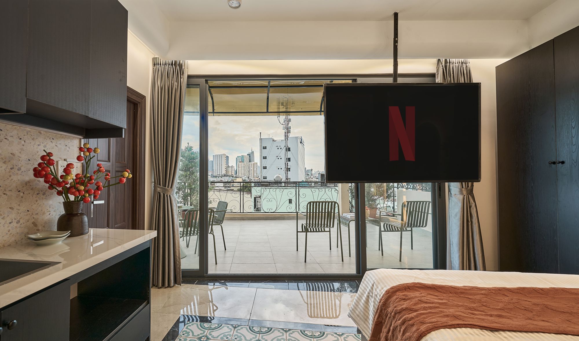 Ein Hotelzimmer mit Bett, einem an der Wand befestigten Fernseher, einem Balkon mit Stühlen und Blick auf die Stadtlandschaft.
