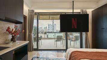 Ein Hotelzimmer mit Bett, einem an der Wand befestigten Fernseher, einem Balkon mit Stühlen und Blick auf die Stadtlandschaft.