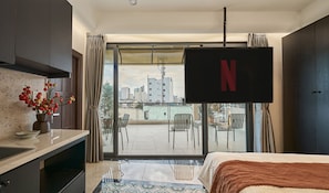 A hotel room with a bed, a TV mounted on the wall, a balcony with chairs, and a view of the cityscape.