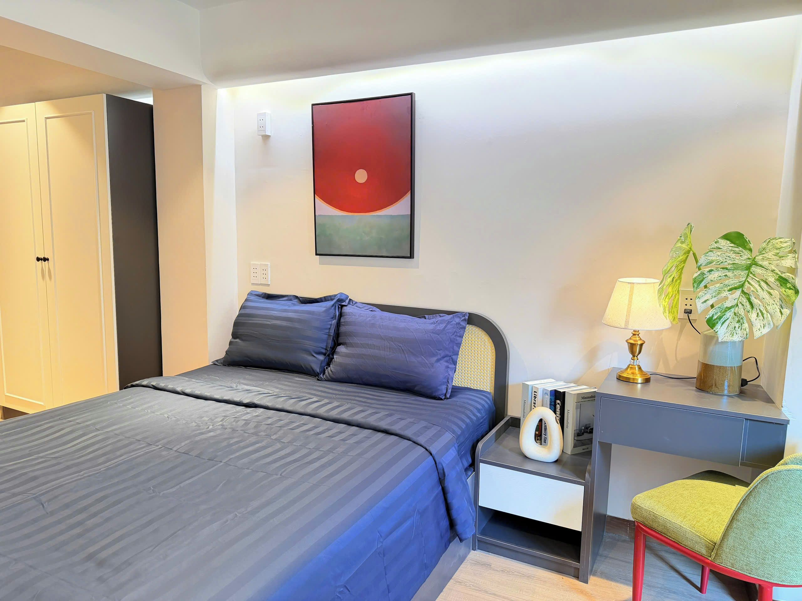 A hotel room with a bed, bedside table with a lamp, a chair, a desk, and a potted plant.