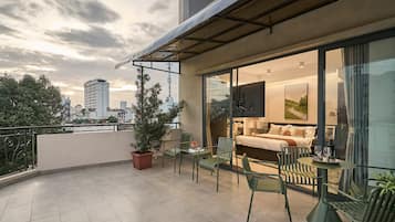 Un balcon aménagé avec un canapé, des chaises et une table, d’où l’on peut admirer une vue sur la ville, avec ses bâtiments et un arbre.