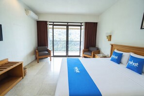 Habitación de hotel con una cama grande, dos sillones, una mesita de madera, una pequeña mesa con un teléfono y vistas a un balcón con cortinas.