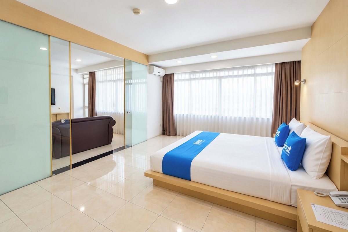 Habitación de hotel moderna con una cama grande, un escritorio con teléfono y una pared de vidrio que separa la habitación de una zona de estar.