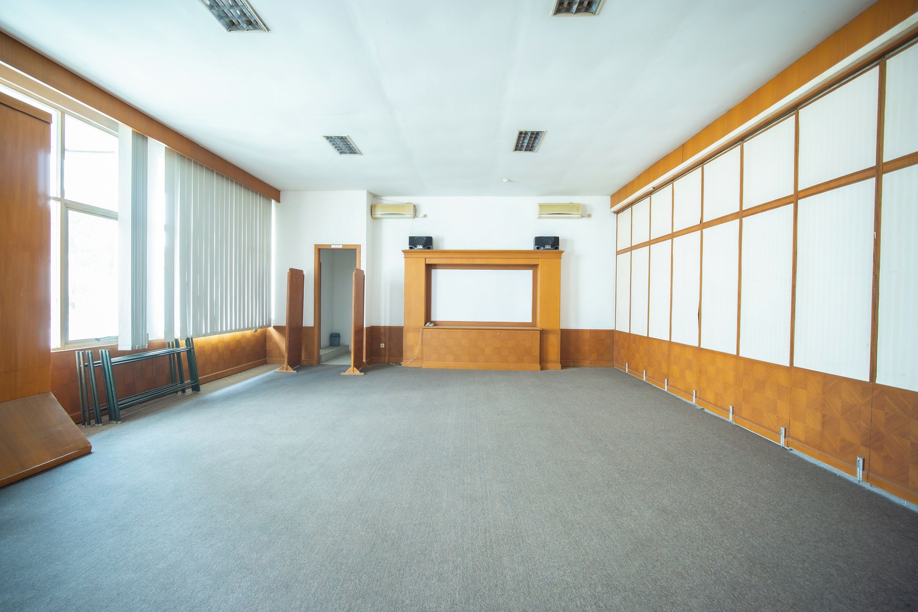 A large, empty room with a carpeted floor, wooden paneling on the walls, a large window on the left, and a door on the right.