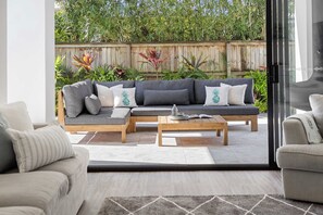 Een moderne buitenruimte met een grijze modulaire bank, een houten salontafel en een glazen deur die uitkomt op een tuin met diverse planten.