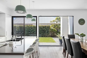 Een moderne keuken met een centraal eiland, een eethoek met stoelen en een uitzicht op een tuin door grote ramen.