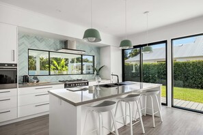 A modern kitchen with a large island, white cabinetry, and a view of a garden through large windows. - Ocean Mist Noosaville Retreat (Noosaville)