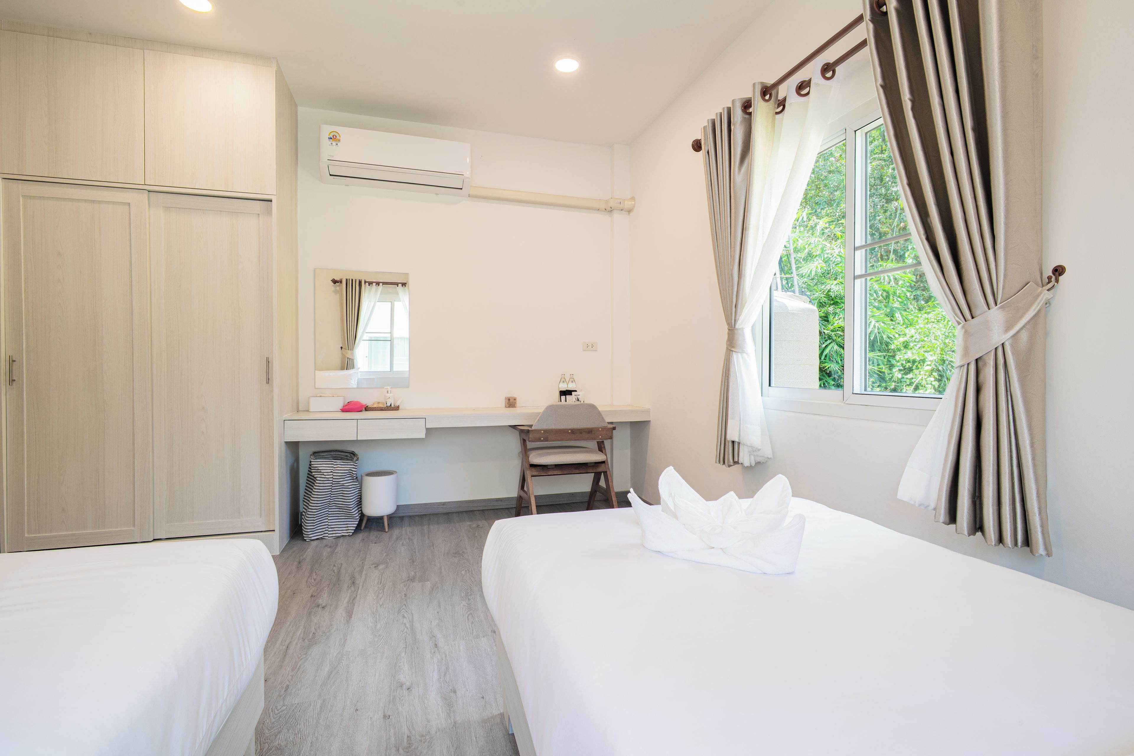 A hotel room with two beds, a desk, a chair, a wardrobe, an air conditioning unit, and a window with curtains.