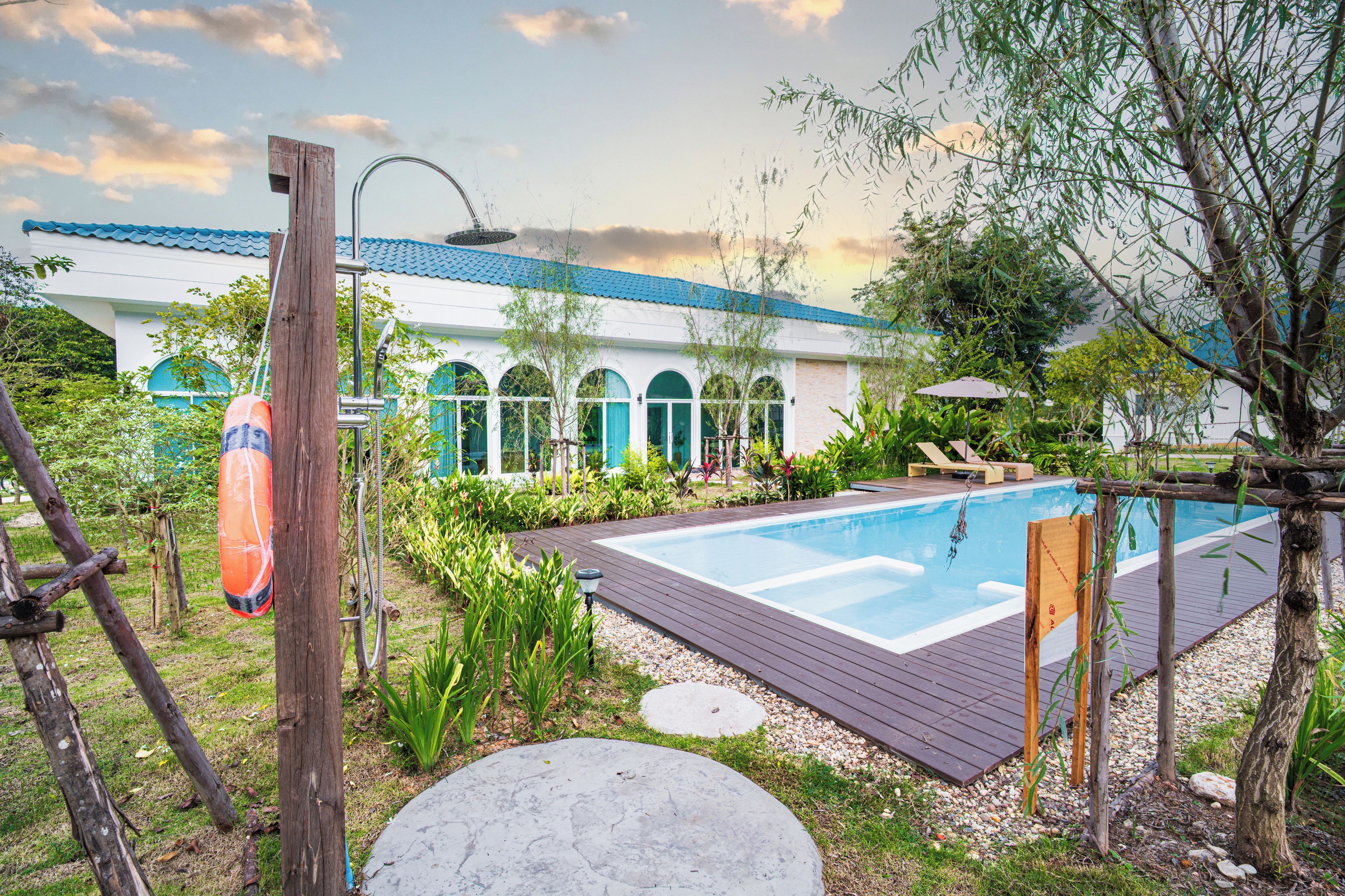 A swimming pool with a wooden deck, surrounded by greenery and a building with a blue roof in the background.