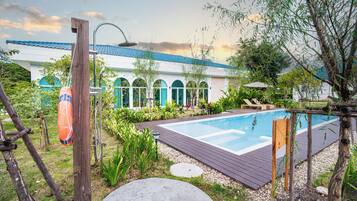 A swimming pool with a wooden deck, surrounded by greenery and a building with a blue roof in the background.