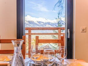 Une table placée devant une fenêtre offrant une vue dégagée sur des montagnes enneigées, avec une rambarde en bois et un vase en verre.