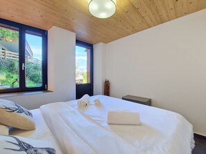 A bedroom with a large bed, two windows, a wooden ceiling, and a view of greenery outside. - Byzance 6 by Interhome (Nendaz)