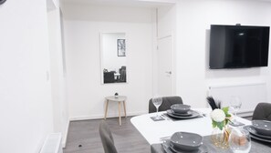 A modern dining area with a white table set for four, a small side table with a plant, and a flat-screen TV mounted on the wall. - Charming Manchester House With 3 Bedrooms (Manchester)