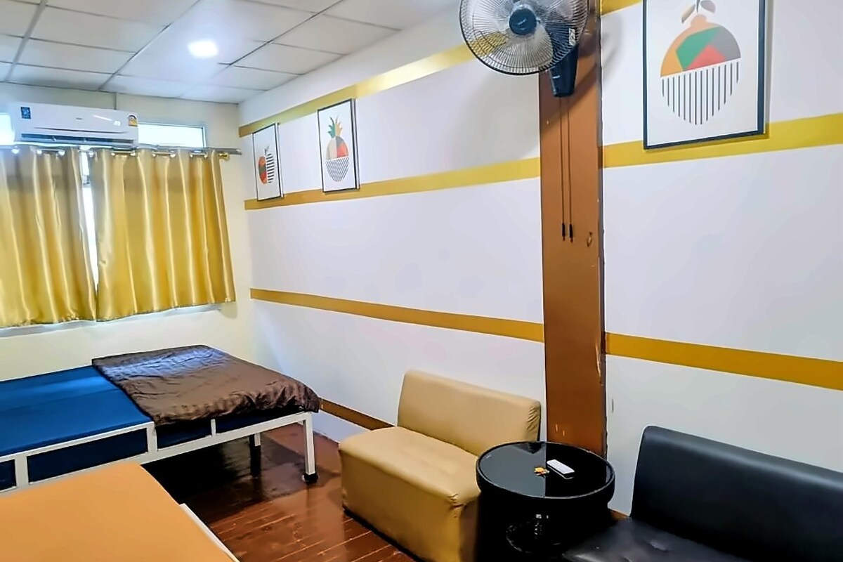 A room with a bed, a sofa, a small table, a fan, and a window with curtains.
