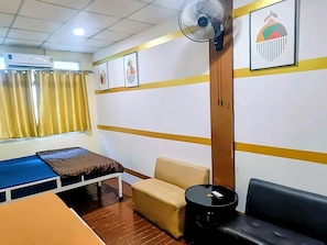 A room with a bed, a sofa, a small table, a fan, and a window with curtains. - The House Hostel 2  (Bangkok)