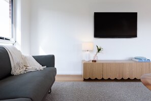 Uma sala de estar moderna com um sofá cinzento, um aparador de madeira, uma TV de tela plana fixada na parede e um abajur sobre o aparador.
