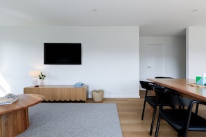 Ein modernes Wohnzimmer mit einem Holzesstisch, schwarzen Stühlen, einem grauen Teppich, einer Holzkonsole, einem flachbildfernseher an der Wand und einer weißen Tür.