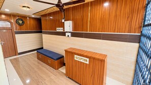 A reception area with a wooden reception desk, a bench, a clock, and a sign indicating check-out time. - Shri Anandam (Vrindavan)