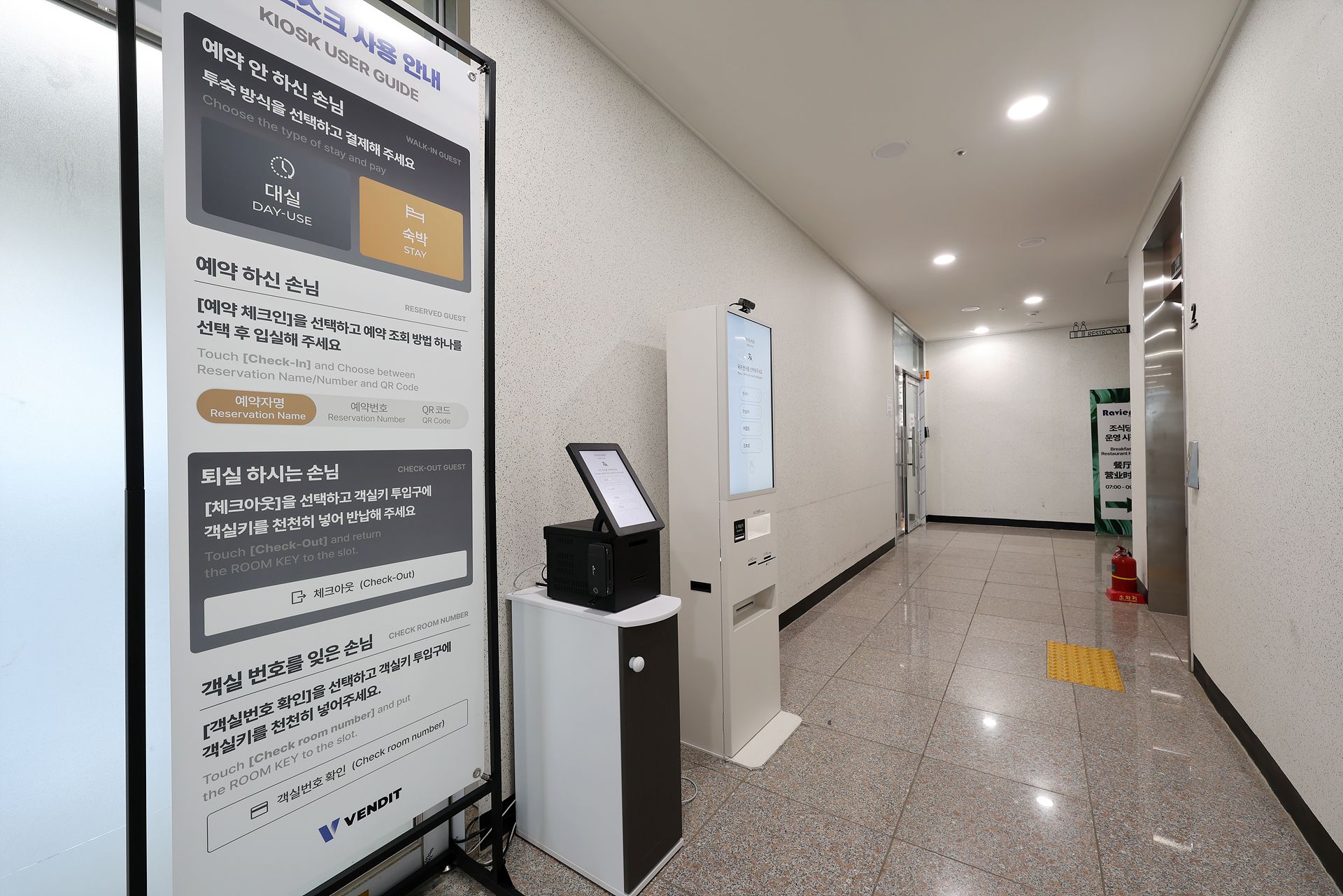 Un pasillo de hotel con un quiosco de registro, una pantalla digital para seleccionar habitaciones y un extintor.
