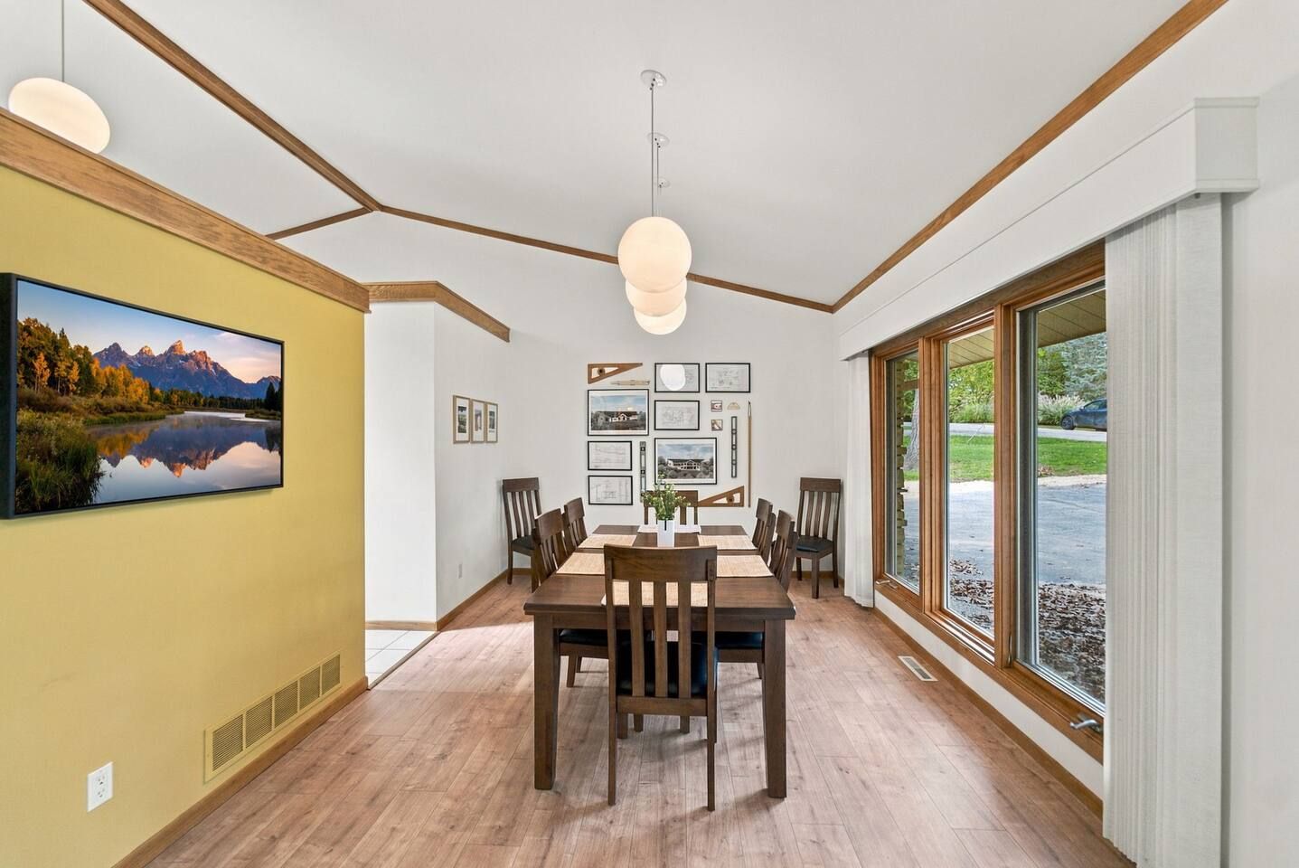 A dining area with a wooden table and chairs, a large window with a view of greenery, and a mounted painting of a mountain landscape.