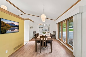 A dining area with a wooden table and chairs, a large window with a view of greenery, and a mounted painting of a mountain landscape. - The Architect’s Overlook | Sweeping Valley Views (Galena)