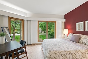 Ein Hotelzimmer mit Bett, Schreibtisch und Stuhl. Große Fenster bieten Blick auf Grünflächen und Häuser.
