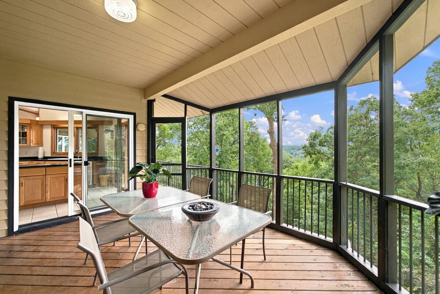 A covered outdoor patio with a table and chairs, a potted plant, and a view of a forested landscape.