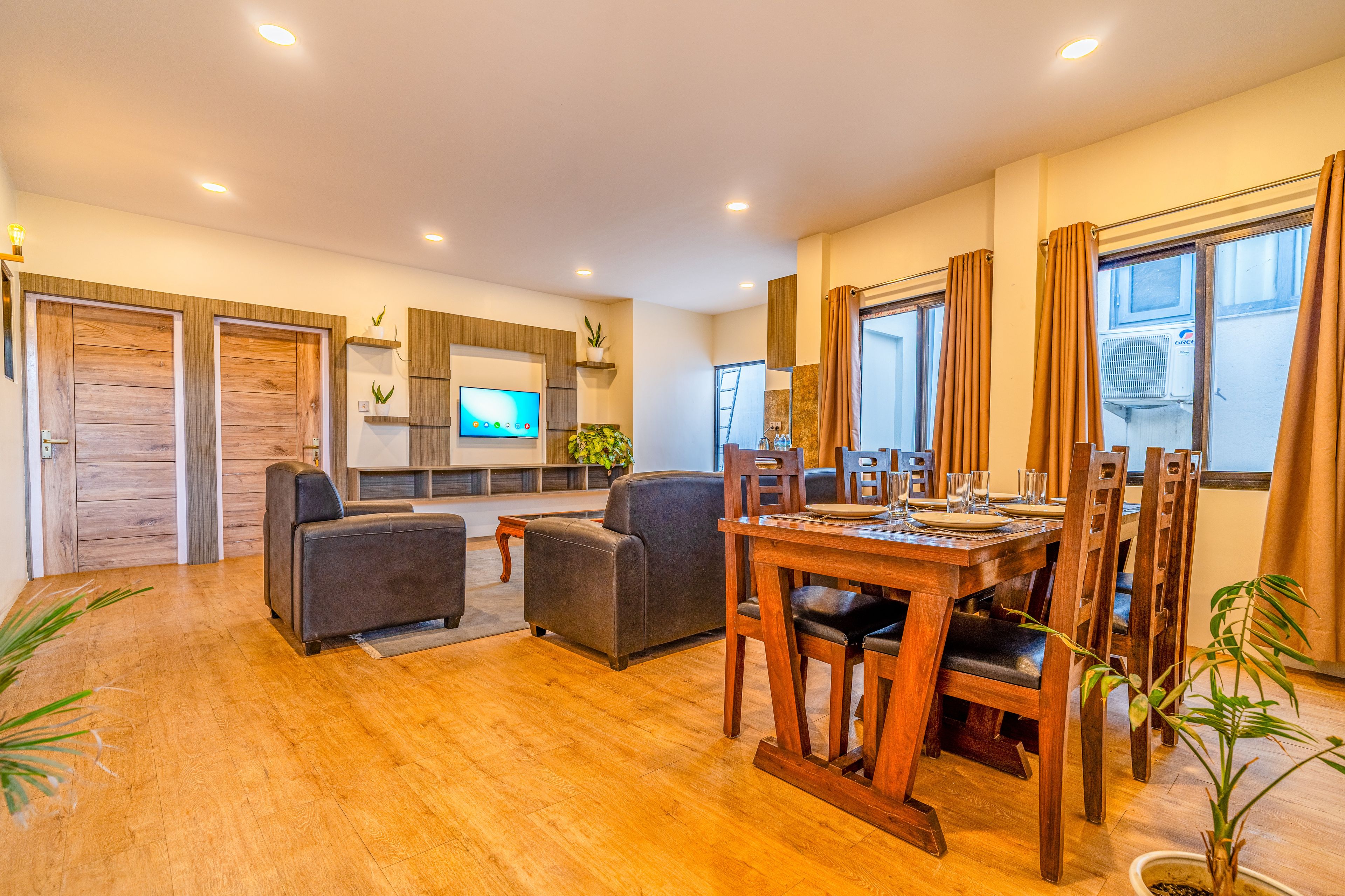 A modern living room with a wooden floor, a dining area with a wooden table and chairs, a flat-screen TV mounted on the wall, and a large window with curtains.