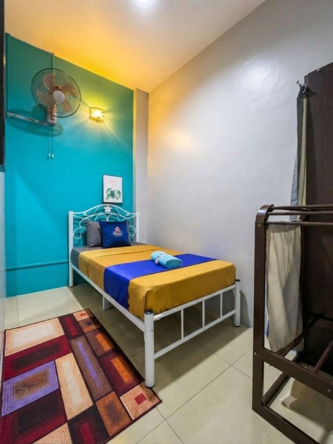 Habitación individual con una pared verde azulado, un ventilador, una alfombra estampada y una ventana con cortinas.
