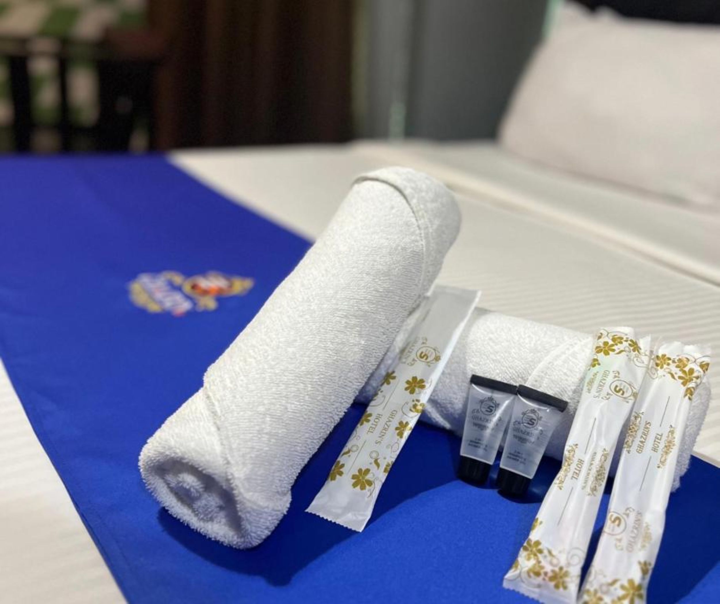 Een net opgemaakt bed met een blauwe spreideken, een opgerolde witte handdoek en een verzorgingsset met een tandenborstel en tandpasta.