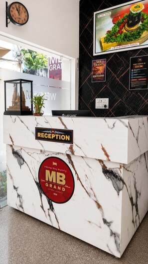 A hotel reception desk with a marble top, a clock, and various signs including a 'MB GRAND' logo. - Hotel MB Grand (Varanasi)