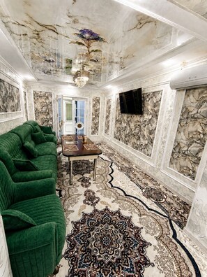 A room with a green sofa, a dining table with a patterned rug, and a stone wall with a mounted TV. - Tranquil 2-bedroom apartment with AC in charming Samarqand (Samarqand)