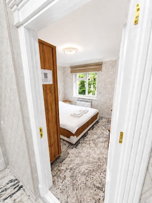 A bedroom with a bed, a window with a view of greenery, a radiator, and a door with a handle.