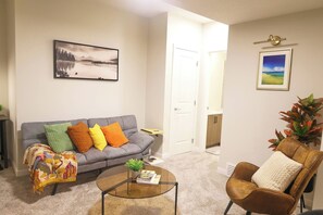 A living room with a grey sofa, colorful cushions, a coffee table with books, a framed landscape painting, and a plant. - Luxury Private Suite, Near Calgary Airport (Calgary)