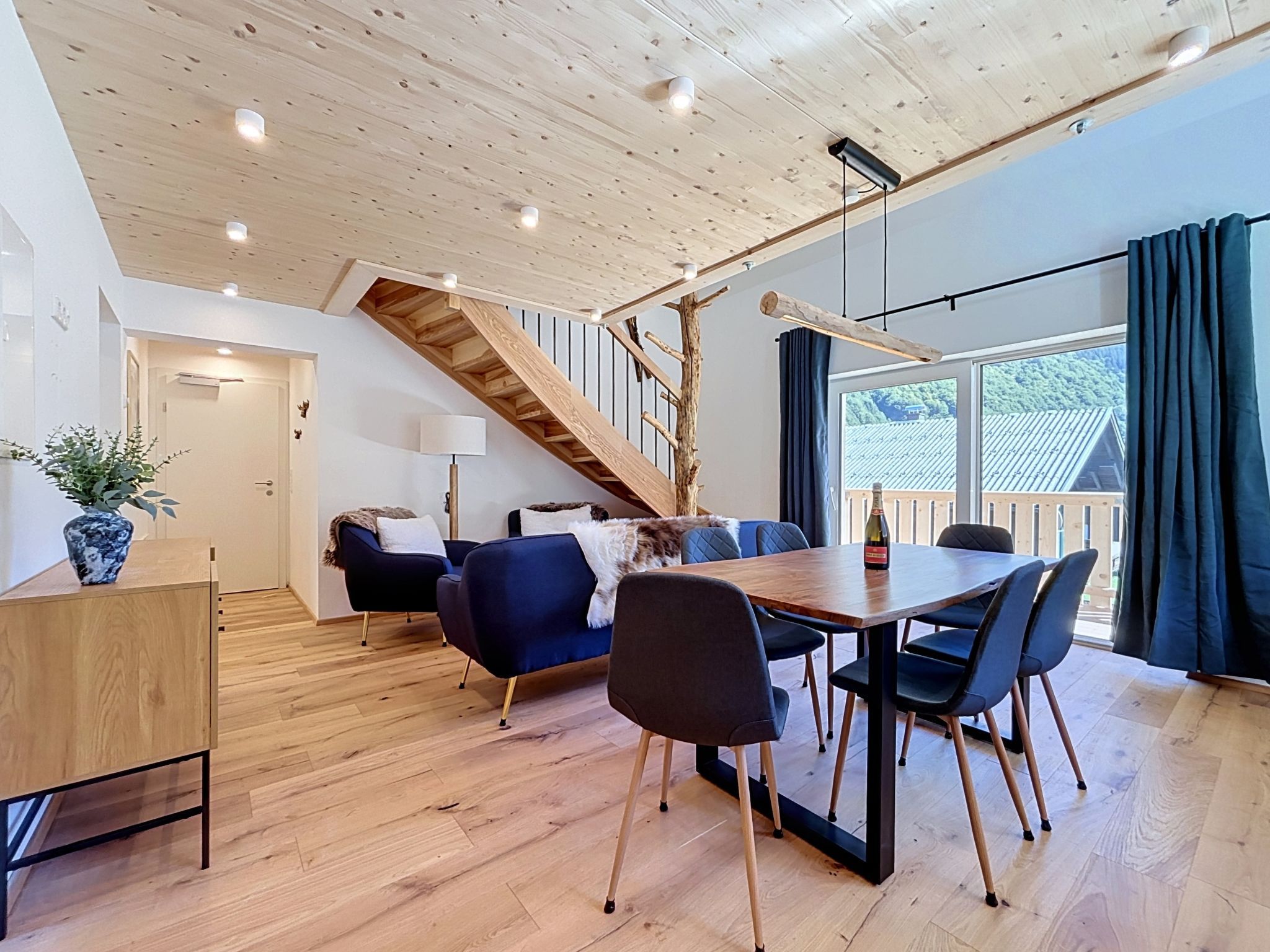 Una habitación amplia con techo de madera, una escalera, un comedor con sillas, una mesa y un gran ventanal con cortinas.
