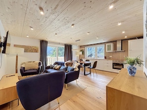 A modern living room with a sofa, a dining table, and a kitchen area with wooden cabinets and a large window.