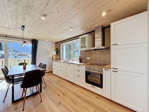 A modern kitchen with wooden flooring, white cabinetry, a dining table with chairs, and a large window offering a view of the outdoors.