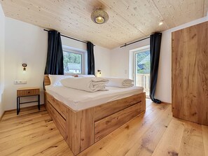 Ein Schlafzimmer mit einem hölzernen Bett, zwei Fenstern mit Vorhängen, einem kleinen Nachttisch und einer Deckenleuchte.