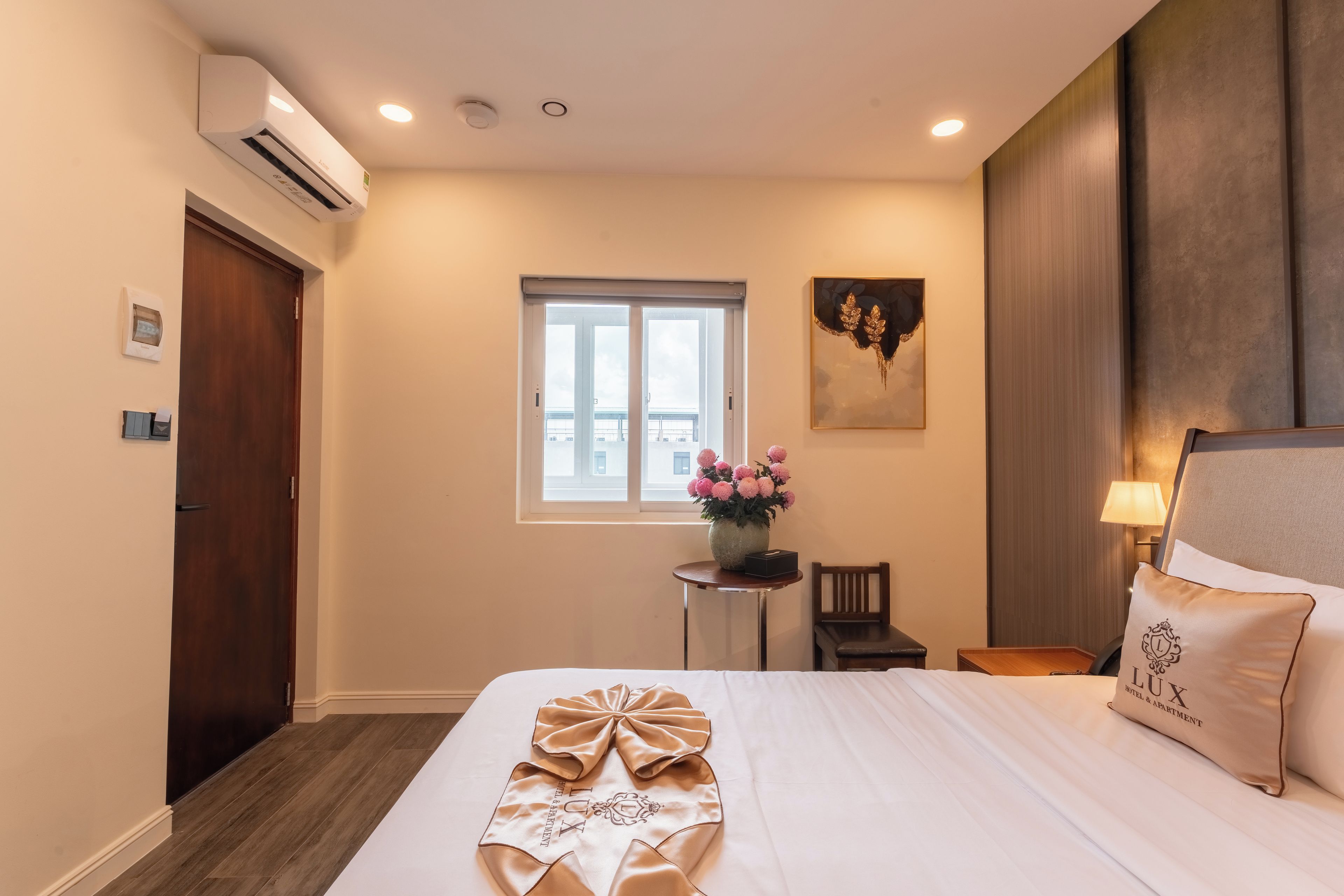 A hotel room with a bed, a window, a small table with a vase of flowers, and a wall-mounted air conditioner.