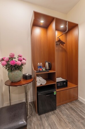 A hotel room with a wooden wardrobe, a small table with a vase of flowers, a black refrigerator, and a bench.