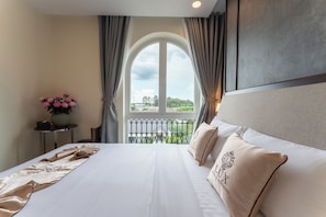 A hotel room with a large bed, a view of greenery through a window, and a small table with a vase of flowers.