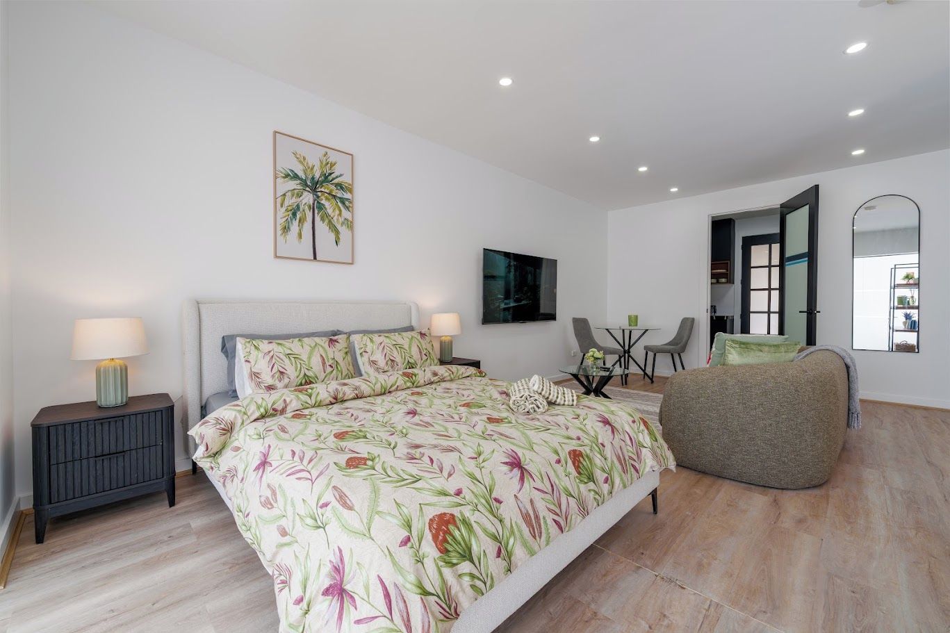 A bedroom with a bed, bedside tables, a TV, a small table with chairs, and a large mirror.