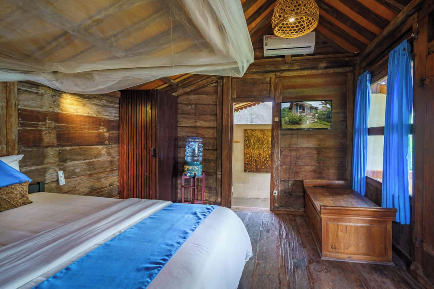A wooden room with a bed, a water dispenser, a bench, and a view of the outside.