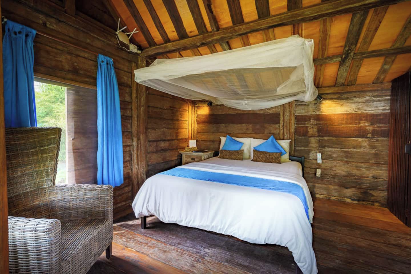 A wooden cabin room with a bed, a wicker chair, and a window with curtains.