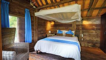 A wooden cabin room with a bed, a wicker chair, and a window with curtains.