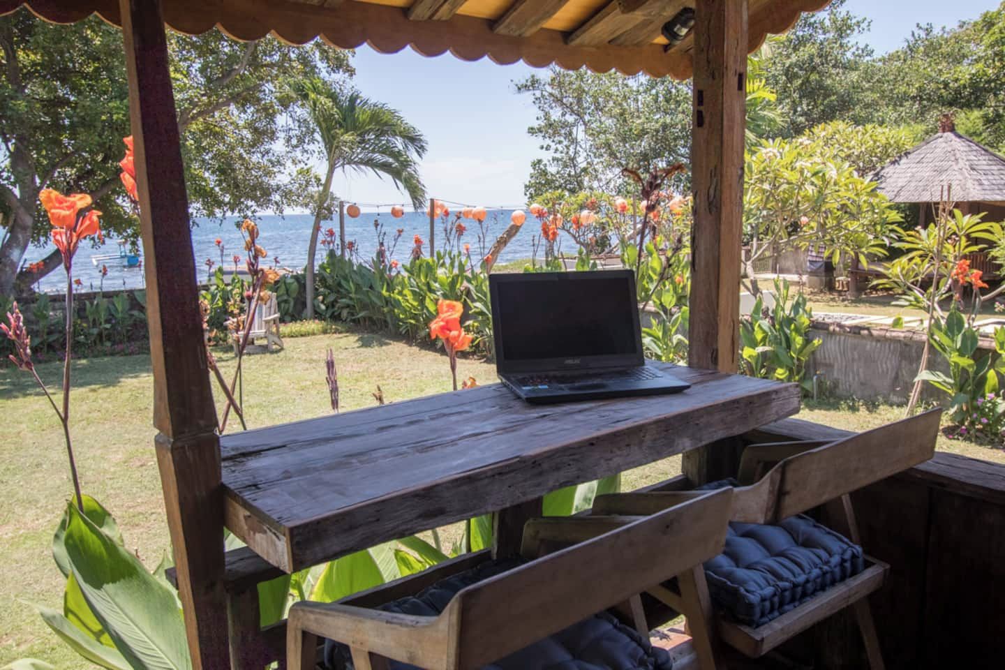 Un tavolo di legno con un portatile, circondato da vegetazione e vista sul mare.